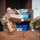 Two U.S. Army soldiers in OCP (Operational Camouflage Pattern) uniforms are sorting and processing mail and packages in what appears to be a military post office. One soldier, a woman wearing glasses, is examining an Amazon package, while a male soldier wearing blue latex gloves handles cardboard boxes. A USPS label reading 'CMR 8 Amentum Unit: 93218' is visible, indicating a military mail facility.