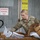 A U.S. Army soldier named Darcy is sorting through a large collection of packages and parcels, including Amazon Prime deliveries, in what appears to be a military mail handling facility. The soldier is wearing OCP (Operational Camouflage Pattern) uniform and is actively working through a pile of mixed packages. The setting is a secured area indicated by an 'Authorized Personnel Only' sign on a corrugated metal wall.