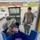 Two U.S. Army soldiers are handling mail or packages at what appears to be a military postal facility, likely at Camp Bondsteel or another KFOR installation in Kosovo. One soldier (name tape reads 'SMITH') is standing and observing while another soldier bends over a large blue bin examining a package. A utility vehicle loaded with boxes is visible in the background near a white KFOR-marked van.