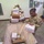 A U.S. Army soldier in MultiCam/OCP camouflage uniform is sorting and handling mail packages in what appears to be a military mail room. The soldier is holding a box from 'Adopt a Platoon,' an organization that supports deployed troops, while surrounded by numerous USPS Priority Mail packages stacked on a cart and in a large canvas mail hamper. The setting appears to be an overseas military installation based on the electrical panel markings showing 220/380 volts.