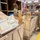 Three U.S. Army soldiers in OCP (Operational Camouflage Pattern) uniforms are sorting and processing mail packages in what appears to be a military post office facility. The packages include USPS Priority Mail boxes and Amazon shipments being organized into large canvas mail carts. An American flag is visible in the background, and the setting resembles a military mail distribution center, possibly overseas given the KFOR patch visible on one soldier's uniform.