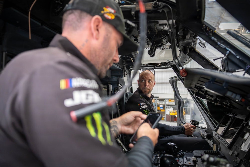 Two individuals wearing NASCAR and Monster Energy branded apparel are inside what appears to be a military aircraft cockpit or simulator. The person in the foreground is looking at a smartphone while the person in the background is seated in the cockpit, looking toward the camera. The setting appears to be an aviation facility or hangar.
