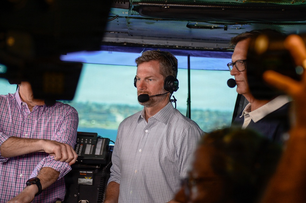 Three men are positioned in what appears to be a military vessel's bridge or control room, with two of them wearing communication headsets. A label visible on equipment reads 'USS CARL VINSON', suggesting this is aboard a U.S. Navy aircraft carrier. The men appear to be observing or broadcasting, possibly in a media or official capacity.