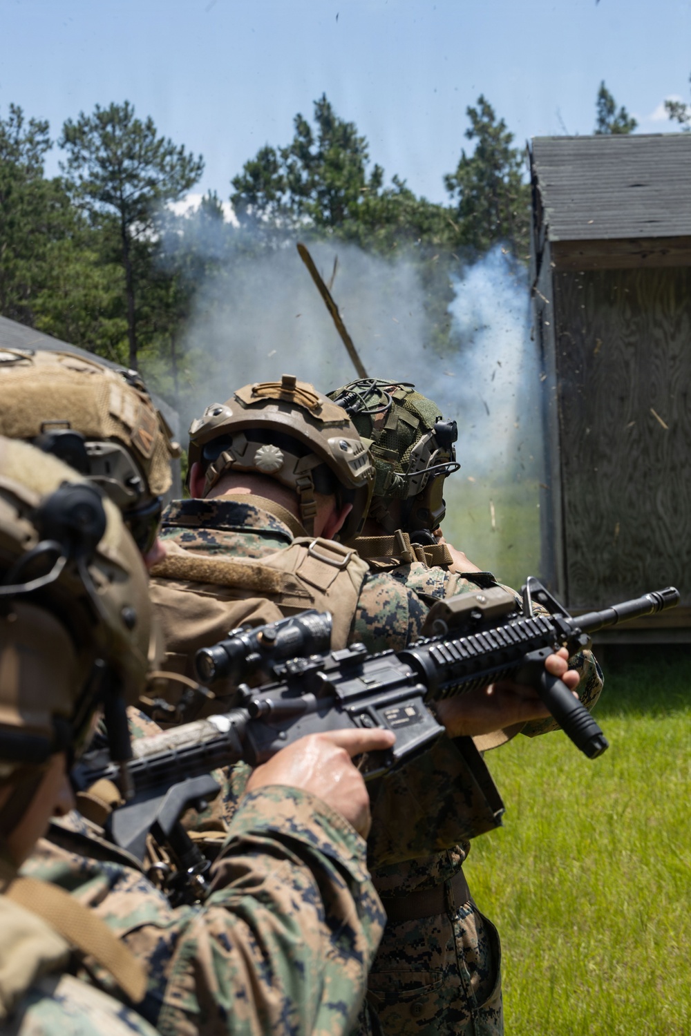 Two military personnel in full combat gear are conducting a breaching or demolition exercise near a structure. Smoke and debris are visible from what appears to be an explosive breach of a wooden building. The personnel are wearing MARPAT (Marine Pattern) digital camouflage uniforms, consistent with U.S. Marine Corps equipment.