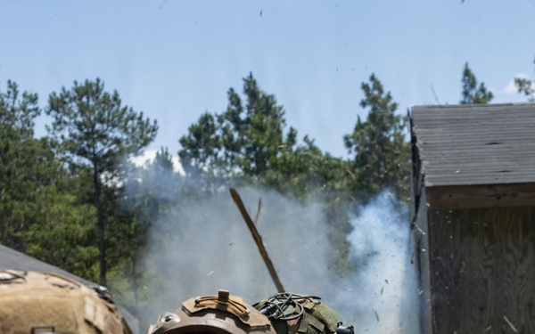 Two military personnel in full combat gear are conducting a breaching or demolition exercise near a structure. Smoke and debris are visible from what appears to be an explosive breach of a wooden building. The personnel are wearing MARPAT (Marine Pattern) digital camouflage uniforms, consistent with U.S. Marine Corps equipment.
