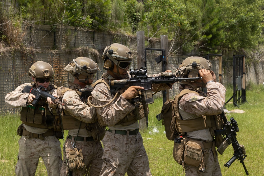 Four military personnel in desert MARPAT camouflage uniforms are conducting a tactical training exercise in an outdoor range environment. The operators are equipped with full combat gear including helmets, plate carriers, and assault rifles, and appear to be practicing a stack or breach formation. The setting includes a constructed training facility with fencing and a doorway frame visible in the background.