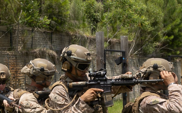 Four military personnel in desert MARPAT camouflage uniforms are conducting a tactical training exercise in an outdoor range environment. The operators are equipped with full combat gear including helmets, plate carriers, and assault rifles, and appear to be practicing a stack or breach formation. The setting includes a constructed training facility with fencing and a doorway frame visible in the background.
