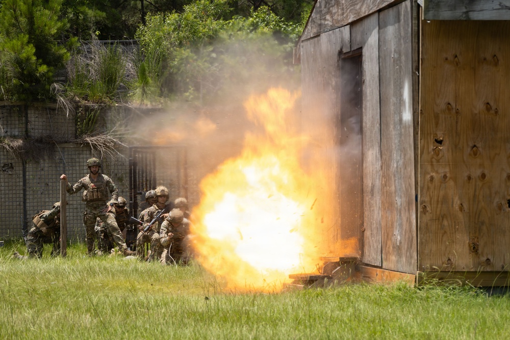 Military personnel in combat uniforms and helmets are taking cover near a wooden structure as a large controlled explosion detonates at its base. The scene appears to be a breaching or demolitions training exercise conducted outdoors in a grassy area with trees in the background. The fireball and smoke indicate the detonation of a breaching charge, likely used to gain entry through a wall or door.