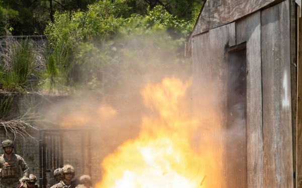 Military personnel in combat uniforms and helmets are taking cover near a wooden structure as a large controlled explosion detonates at its base. The scene appears to be a breaching or demolitions training exercise conducted outdoors in a grassy area with trees in the background. The fireball and smoke indicate the detonation of a breaching charge, likely used to gain entry through a wall or door.