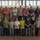 A group of approximately 19 men posed for a formal group photo inside an industrial facility or workshop. They are arranged in two rows — a back row elevated on a platform and a front row standing on the floor — in front of a large tan/beige shipping container or industrial enclosure. The individuals appear to be workers, tradespeople, or trainees based on their attire and the industrial setting.