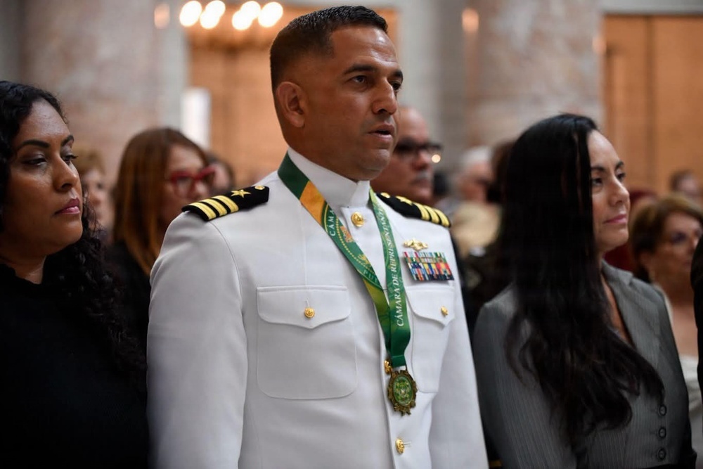 A military officer in a white dress uniform is standing at what appears to be a formal ceremony, wearing a green and gold medallion sash inscribed with 'Cámara de Representantes.' He is adorned with multiple service ribbons and rank insignia consistent with a senior naval officer. He is flanked by two women in formal attire in what appears to be an official government or ceremonial hall.