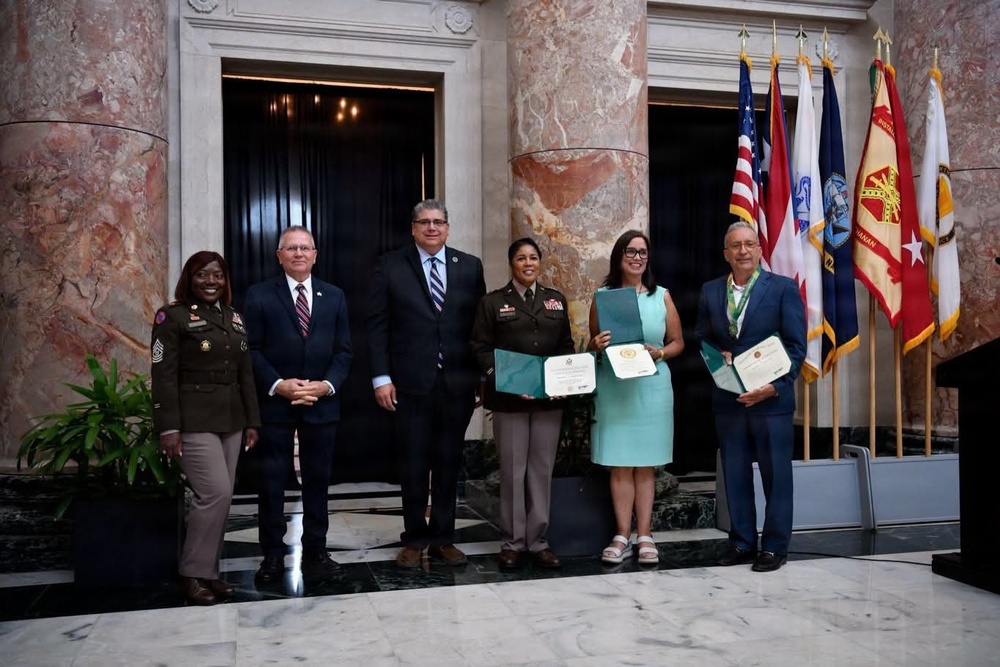 A formal military awards ceremony taking place in an ornate marble building, likely a government or military headquarters. Six individuals are posed for a group photo, with three of them holding certificates or award documents. Two individuals are in U.S. Army uniforms while the others are in civilian attire.