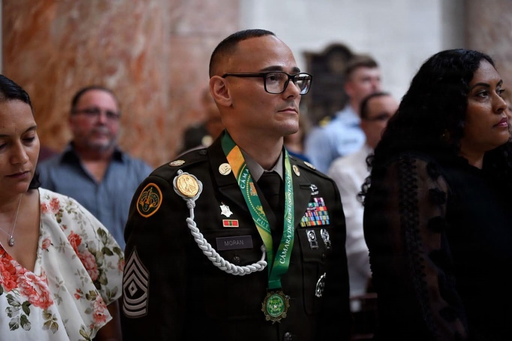 A U.S. Army soldier in dress green uniform, identified by name tape as 'MORAN' with a Sergeant Major rank insignia, stands at attention during what appears to be a formal ceremony. He is wearing a green and gold medal sash reading 'CÁMARA DE REPRESENTANTES' (Chamber of Representatives), along with multiple service ribbons and decorations. He is flanked by two civilian women in an ornate marble-walled venue.