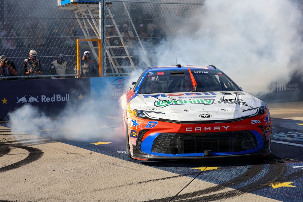 A NASCAR Toyota Camry race car numbered 67, driven by 'Heim', performs a burnout celebration on pit road, generating large clouds of tire smoke. The car features sponsorships from Mobil 1, O'Reilly Auto Parts, and 23XI Racing livery in red, white, and blue colors. The event appears to be at a NASCAR race venue with Red Bull branding visible on the pit wall barrier.