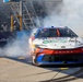 A NASCAR Toyota Camry race car numbered 67, driven by 'Heim', performs a burnout celebration on pit road, generating large clouds of tire smoke. The car features sponsorships from Mobil 1, O'Reilly Auto Parts, and 23XI Racing livery in red, white, and blue colors. The event appears to be at a NASCAR race venue with Red Bull branding visible on the pit wall barrier.