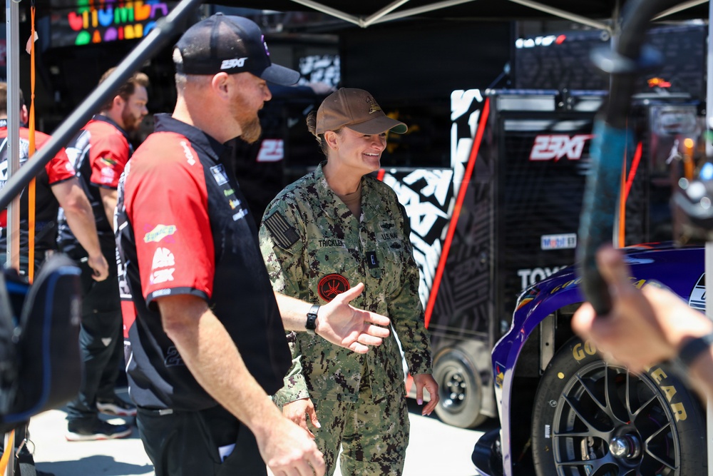 A U.S. Navy service member wearing NWU Type III camouflage uniform with a name tape reading 'THICKLER' is conversing and gesturing with a NASCAR pit crew member in a red 23XI Racing team uniform. The setting is a NASCAR garage/pit area with 23XI Racing team equipment, a race car with Goodyear tires, and additional crew members visible in the background. The interaction appears to be a military appreciation or promotional visit to a NASCAR event.