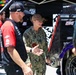 A U.S. Navy service member wearing NWU Type III camouflage uniform with a name tape reading 'THICKLER' is conversing and gesturing with a NASCAR pit crew member in a red 23XI Racing team uniform. The setting is a NASCAR garage/pit area with 23XI Racing team equipment, a race car with Goodyear tires, and additional crew members visible in the background. The interaction appears to be a military appreciation or promotional visit to a NASCAR event.