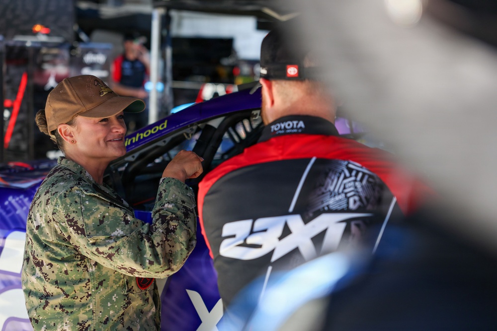 A person in military camouflage uniform and tan cap is conversing with an individual wearing a 23XI Racing team jacket with Toyota branding. The setting appears to be a NASCAR pit or garage area, with a purple/blue race car visible in the background. The interaction appears friendly and engaged.