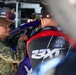 A person in military camouflage uniform and tan cap is conversing with an individual wearing a 23XI Racing team jacket with Toyota branding. The setting appears to be a NASCAR pit or garage area, with a purple/blue race car visible in the background. The interaction appears friendly and engaged.