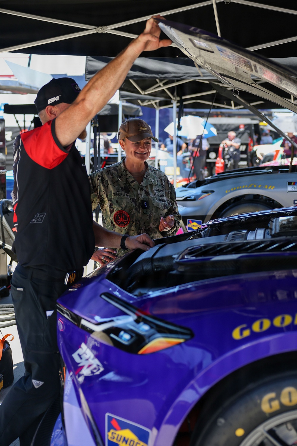 A U.S. Navy service member in camouflage uniform is receiving a tour or explanation of a NASCAR race car in the pit/garage area. A team member wearing a '23XI Racing' cap is lifting the hood/panel of a purple NASCAR vehicle. The setting appears to be a NASCAR Cup Series event based on visible branding.