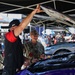 A U.S. Navy service member in camouflage uniform is receiving a tour or explanation of a NASCAR race car in the pit/garage area. A team member wearing a '23XI Racing' cap is lifting the hood/panel of a purple NASCAR vehicle. The setting appears to be a NASCAR Cup Series event based on visible branding.