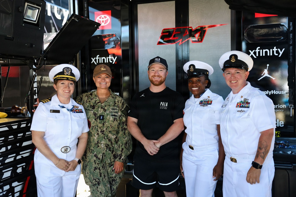 Five individuals pose for a group photo in front of what appears to be a NASCAR racing team hauler or pit area setup for the 23XI Racing team. Four of the individuals are wearing U.S. Navy uniforms — three in white dress uniforms and one in digital camouflage (NWU Type I) — while the fifth person in the center is a civilian male wearing a black 'Pala Casino' shirt and Jordan brand shorts. The setting features visible sponsorship branding including Xfinity, Toyota, and 23XI Racing logos.