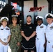 Five individuals pose for a group photo in front of what appears to be a NASCAR racing team hauler or pit area setup for the 23XI Racing team. Four of the individuals are wearing U.S. Navy uniforms — three in white dress uniforms and one in digital camouflage (NWU Type I) — while the fifth person in the center is a civilian male wearing a black 'Pala Casino' shirt and Jordan brand shorts. The setting features visible sponsorship branding including Xfinity, Toyota, and 23XI Racing logos.