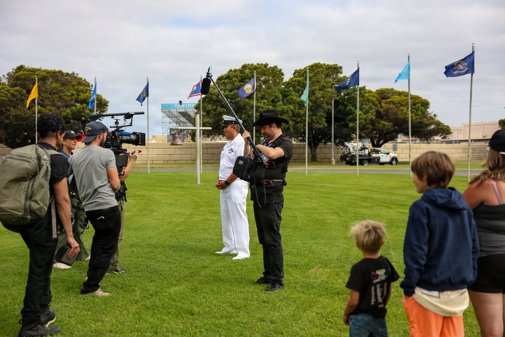 A film or news crew is conducting an outdoor interview with a naval officer in white dress uniform on a green lawn. The scene appears to take place at a military installation, evidenced by multiple state and organizational flags visible on flagpoles in the background. Bystanders, including children and adults, observe the filming from the right side of the frame.