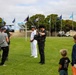 A film or news crew is conducting an outdoor interview with a naval officer in white dress uniform on a green lawn. The scene appears to take place at a military installation, evidenced by multiple state and organizational flags visible on flagpoles in the background. Bystanders, including children and adults, observe the filming from the right side of the frame.