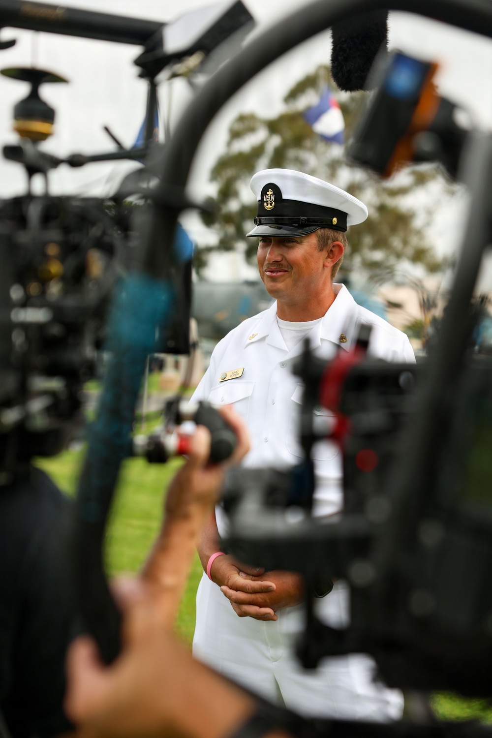 A U.S. Navy enlisted sailor in white dress uniform is being filmed or interviewed outdoors, likely at a public event or media engagement. The sailor wears a white combination cover with 'USN' insignia and a chief petty officer anchor device, suggesting senior enlisted rank. A camera operator in the foreground holds a gimbal-stabilized camera rig, with a boom microphone visible at the top of the frame.