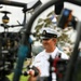 A U.S. Navy enlisted sailor in white dress uniform is being filmed or interviewed outdoors, likely at a public event or media engagement. The sailor wears a white combination cover with 'USN' insignia and a chief petty officer anchor device, suggesting senior enlisted rank. A camera operator in the foreground holds a gimbal-stabilized camera rig, with a boom microphone visible at the top of the frame.