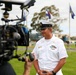 A U.S. Navy enlisted sailor in white dress uniform is being filmed by a professional camera crew outdoors on what appears to be a military base. The sailor is wearing a combination cover (white cap) with gold insignia and has ribbons and badges on his uniform. In the background, military flags are visible on flagpoles along with what appears to be a military helicopter on display.