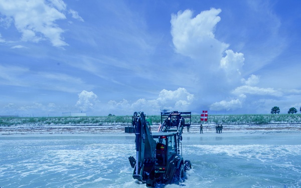 A military or amphibious landing operation is underway, with a backhoe/excavator vehicle driving off the ramp of a landing craft into shallow coastal waters toward a beach. The image is taken from the perspective of the vessel's open bow ramp, showing the vehicle mid-disembarkation in churning surf. Personnel and military vehicles are visible on the shoreline in the background.