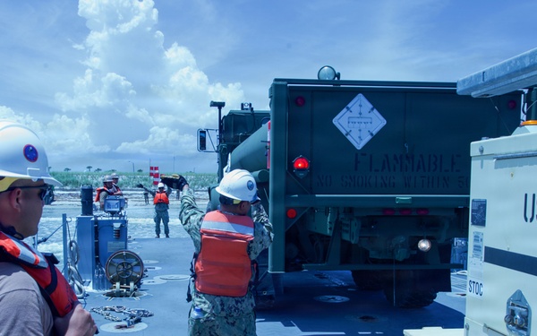 U.S. Navy personnel are conducting a fuel transfer or logistics operation on what appears to be a landing craft or ferry deck near a shoreline. A military fuel truck marked 'FLAMMABLE' is positioned on the deck alongside equipment labeled 'USN 51-288' and marked for 'JP-8 OR DL-2 FUEL'. Personnel wearing white hard hats and orange life vests are directing and overseeing the operation.