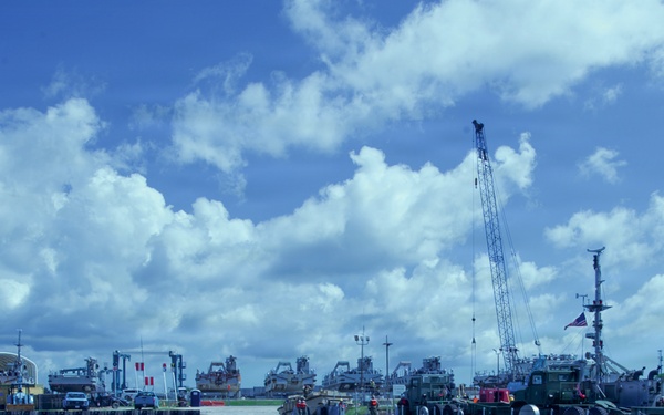 A military or industrial port facility photographed from the water, showing a floating causeway or pontoon bridge system with vessels and heavy equipment. A large crane is prominently visible on the right side, and multiple large ships are docked in the background. The scene suggests active logistics or engineering operations at a naval or military installation.