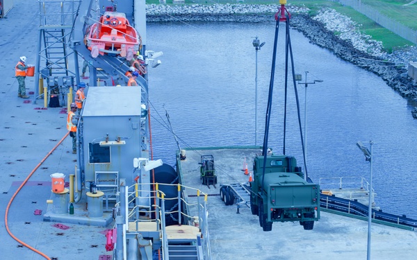 An aerial view of a military or commercial vessel docked at a port facility, with personnel in high-visibility safety gear conducting loading or unloading operations. A military truck (appears to be an LMTV or similar tactical vehicle) is positioned on the dock near a crane or lifting apparatus. Multiple workers are visible on both the ship deck and the dock, coordinating what appears to be a vehicle offload or onload operation.