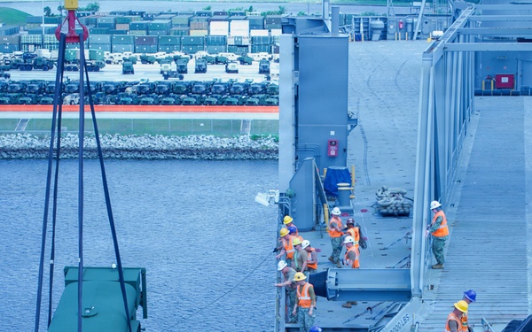 A military logistics operation is underway at a port facility, where a green military tactical vehicle is being lifted by crane from or onto a large vessel. Multiple workers wearing orange safety vests and hard hats are stationed along the ship's deck, coordinating the lift operation. In the background, a staging area on shore contains numerous military vehicles parked in organized rows.