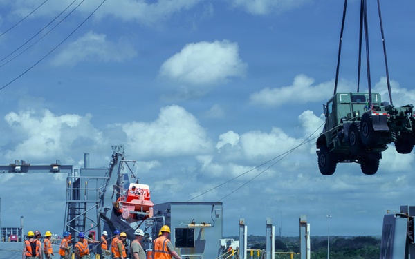 Military personnel in orange safety vests and hard hats are conducting a cargo loading or unloading operation on what appears to be a large military or cargo vessel at a port facility. A military truck (olive drab, likely a medium tactical vehicle) is being lifted by crane rigging and suspended in the air. The scene depicts a coordinated logistics operation with multiple workers managing lines and overseeing the lift.
