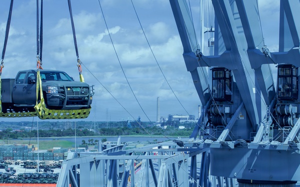 A military or port loading operation is underway at a naval facility, where a dark-colored pickup truck is being lifted by crane using yellow cargo straps onto or off of a large gray military vessel. Multiple workers in high-visibility safety gear and hard hats are visible on the ship's deck and ramp structure. In the background, a staging area contains numerous military vehicles including HMMWVs and other equipment.