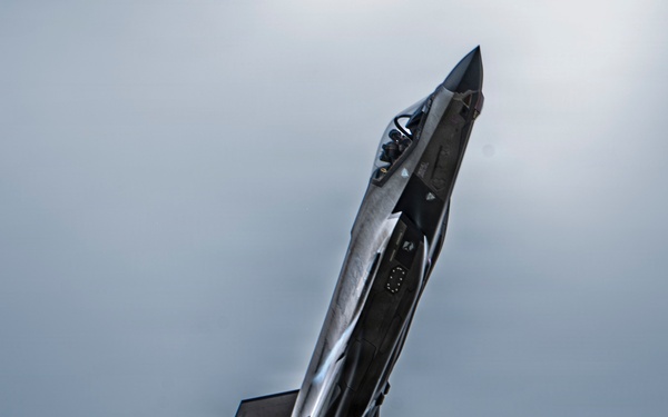 A fifth-generation stealth fighter jet, consistent with an F-35 Lightning II, is captured in a near-vertical climb maneuver against an overcast sky. The aircraft is photographed from below at a dramatic angle, showcasing its underside and delta-wing configuration. Condensation trails are visible streaming from the aircraft's exhaust nozzle.