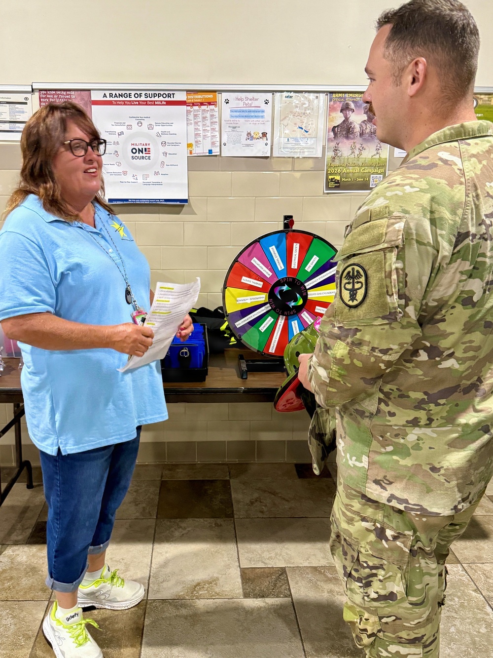 A civilian woman in a light blue polo shirt is conversing with a U.S. Army soldier in MultiCam (OCP) uniform at what appears to be a military information or outreach event. Behind them is a colorful 'Spin to Win' prize wheel and several informational posters on the wall, including a Military OneSource 'A Range of Support' poster and an Army Emergency Relief 2026 Annual Campaign poster. The setting appears to be inside a military installation building.