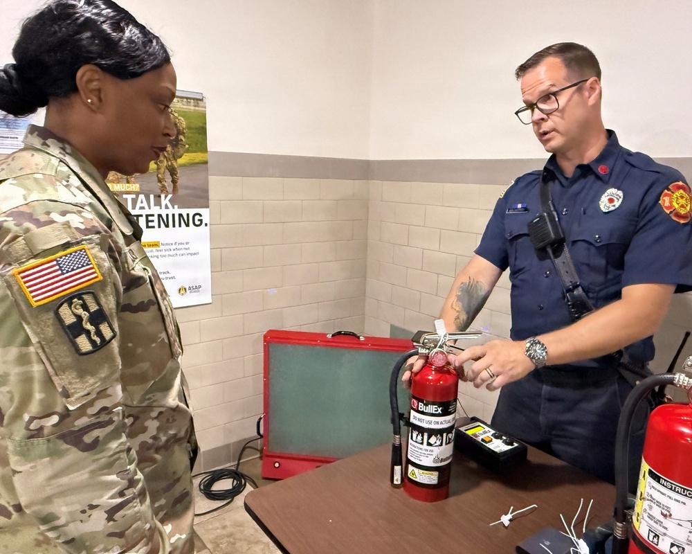 A firefighter/fire safety instructor is demonstrating a BullEx training fire extinguisher to a U.S. Army soldier in a military facility. The firefighter, wearing a navy blue uniform with a fire department patch, is explaining the extinguisher's components while the soldier in Army Combat Uniform (ACU) with an American flag patch observes attentively. The setting appears to be a fire safety training session.