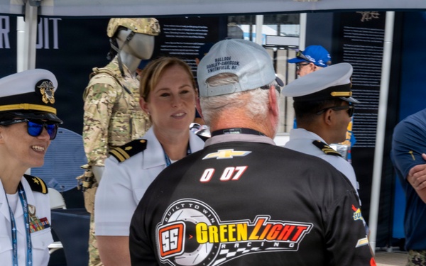 The image shows a Navy recruitment or outreach booth at what appears to be a NASCAR or motorsports event. Military personnel in white dress uniforms and camouflage are interacting with a civilian wearing a 'SS GreenLight Racing' team shirt numbered '007'. The setting includes a display tent with military equipment, uniforms on mannequins, and Navy branding.