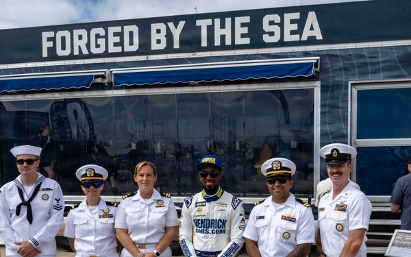 A NASCAR driver wearing a Hendrick Cars racing suit poses with five U.S. Navy personnel in white dress uniforms in front of a Navy recruitment display trailer. The trailer features the slogan 'Forged by the Sea' and promotes Navy.com, along with an 'All Hands' emblem. The setting appears to be a NASCAR race event where the Navy has set up a promotional exhibit.