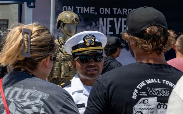 A U.S. Navy officer in dress whites and cover (hat) is speaking with a group of civilians at what appears to be a public military recruitment or outreach event. A recruitment banner reading 'ON TIME, ON TARG... NEVE...' is visible in the background along with a display of military tactical gear. The crowd includes several young adults in casual clothing, suggesting a public event such as an air show or fleet week.