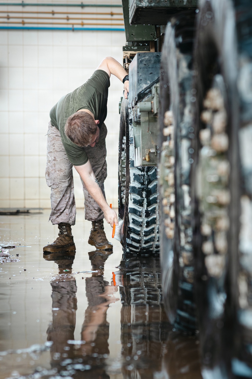 A military service member in camouflage utilities and olive drab t-shirt is scrubbing the track and road wheels of a tracked military vehicle using an orange-handled brush. The work is being performed on a wet floor inside what appears to be a maintenance bay or motor pool facility. The image captures routine post-operation vehicle maintenance, with soapy water and debris visible on the floor.