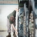 A military service member in camouflage utilities and olive drab t-shirt is scrubbing the track and road wheels of a tracked military vehicle using an orange-handled brush. The work is being performed on a wet floor inside what appears to be a maintenance bay or motor pool facility. The image captures routine post-operation vehicle maintenance, with soapy water and debris visible on the floor.