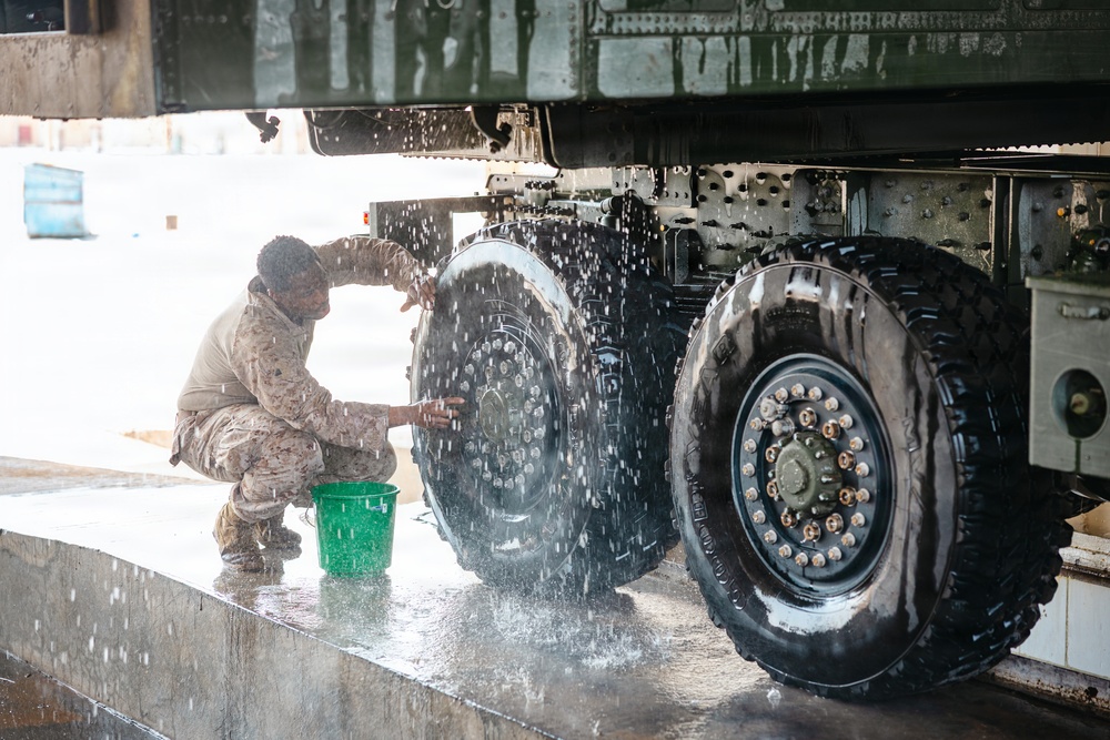 A military service member in camouflage uniform is manually washing the large tires and undercarriage of a military vehicle. The soldier is crouched on a wet concrete wash platform, using a green bucket and hands to scrub the dual rear tires. Water and soap suds are visibly splashing around the work area.
