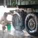 A military service member in camouflage uniform is manually washing the large tires and undercarriage of a military vehicle. The soldier is crouched on a wet concrete wash platform, using a green bucket and hands to scrub the dual rear tires. Water and soap suds are visibly splashing around the work area.
