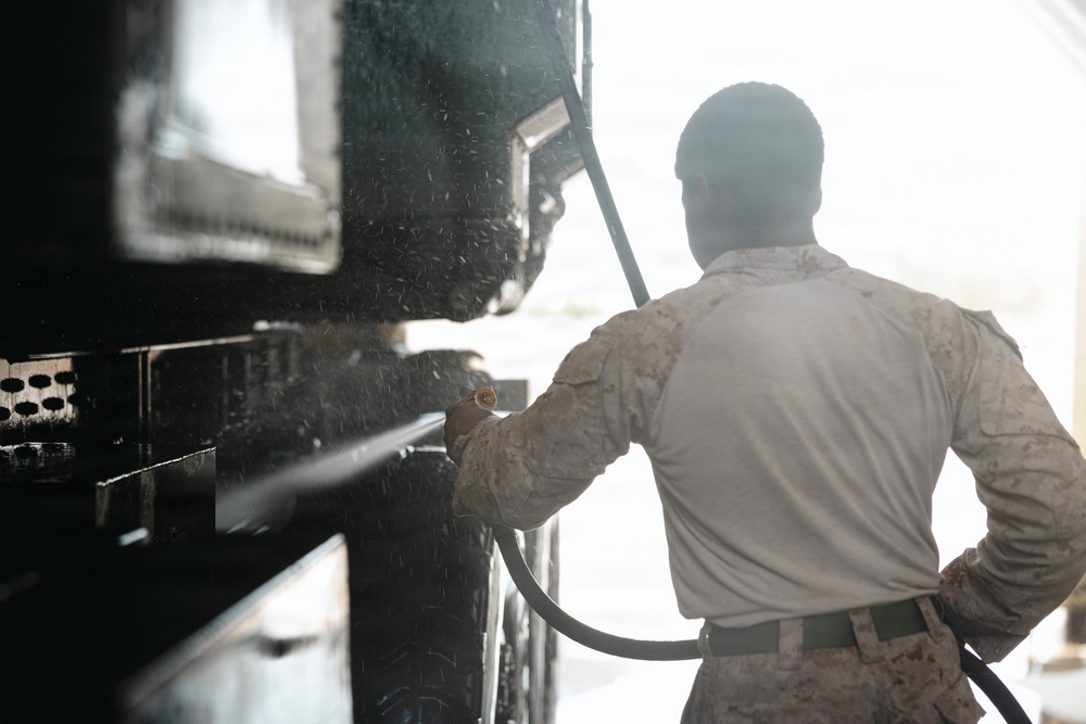 A person wearing military camouflage uniform is using a pressure washer to clean a large dark-colored military vehicle or equipment. The image is taken from behind the individual, with strong backlighting creating a silhouette effect. Water spray and debris are visibly dispersing from the point of contact with the surface being cleaned.